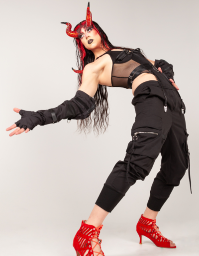 Saltina Shaker wearing a black bodysuit, black gloves, and red horns. She's posing with her back arched dramatically, reaching and looking to her right.