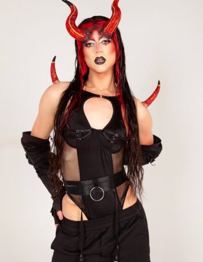 Saltina Shaker wearing a black bodysuit, black gloves, and red horns looking at the camera.