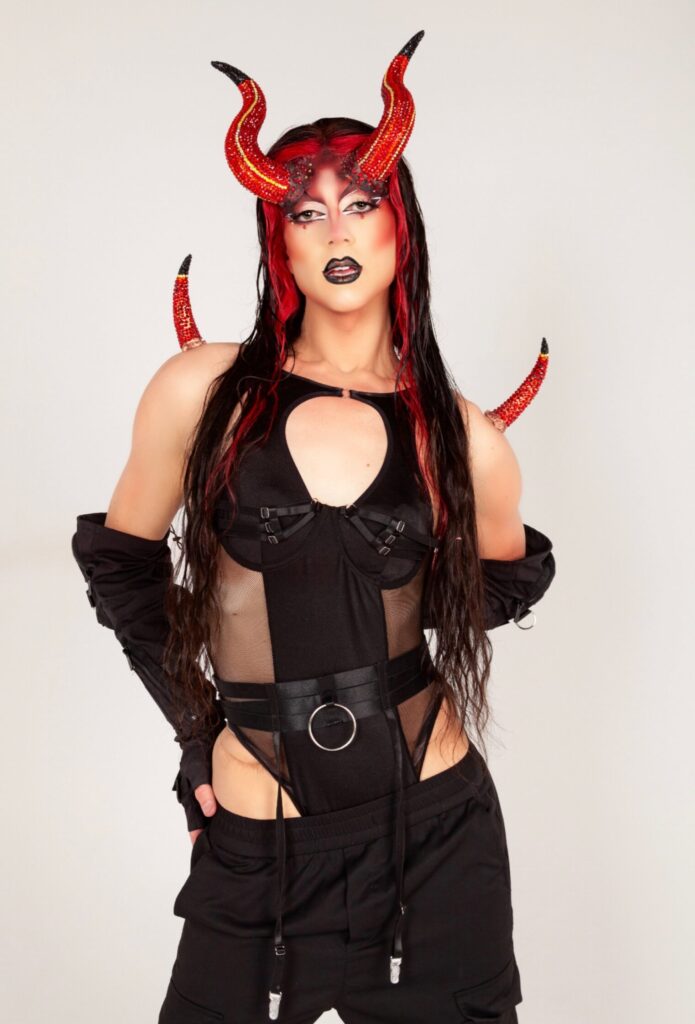 Saltina Shaker wearing a black bodysuit, black gloves, and red horns looking at the camera.