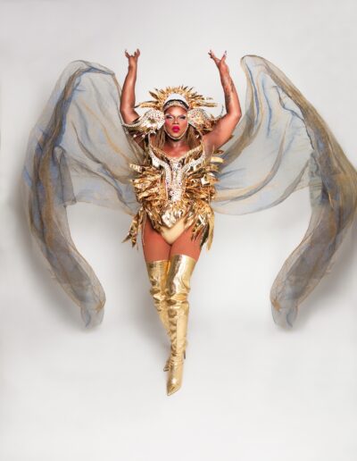 Aimee Yonce Shennel in a golden sun-inspired outfit with matching headdress and silver sheer cape, throwing the cape in the air to look like wings.