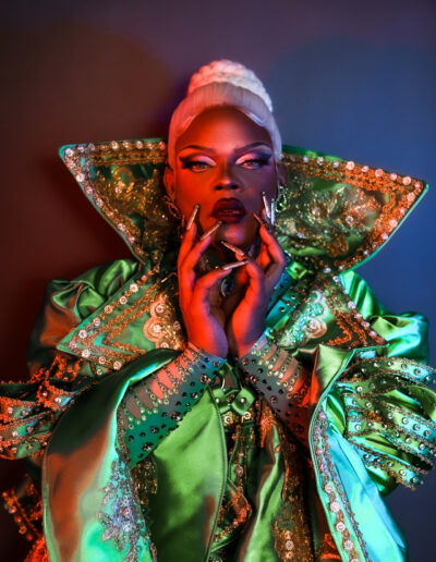 Bust-up view of Aimee Yonce Shennel wearing an emerald green coat with gold embellishments, staring dramatically into the camera.