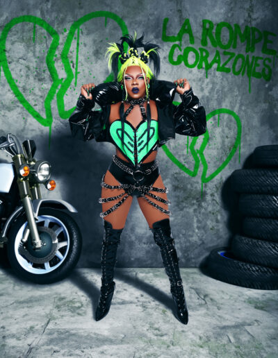 Aimee Yonce Shennel wearing a black and green biker-inspired outfit with black jacket, standing in front of a motorcycle and some tires.