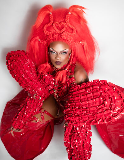 Top view of Aimee Yonce Shennel kneeling in a red strappy bodysuit with dramatic sleeves and matching cape.