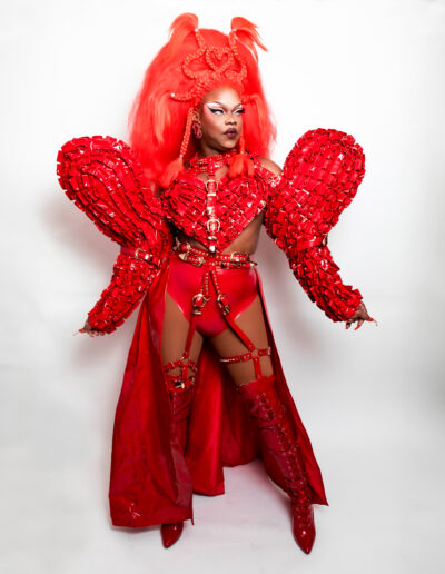 Aimee Yonce Shennel wearing a red strappy bodysuit with dramatic sleeves and matching cape, looking to her left.