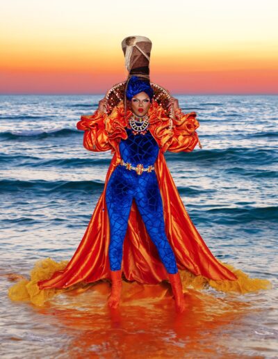 Aimee Yonce Shennel wearing a blue velvet bodysuit and bright oragnge cape, posing in front of an ocean sunset backdrop.