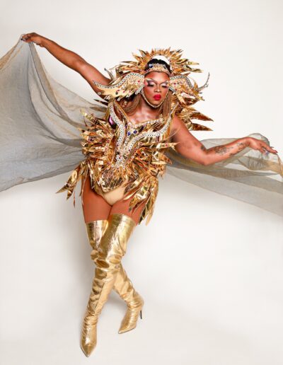 Aimee Yonce Shennel posing like a dancer in a golden sun-inspired outfit with matching headdress and silver sheer cape.