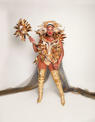 Aimee Yonce Shennel in a golden sun-inspired outfit with matching headdress and silver sheer cape, holding a matching scepter.