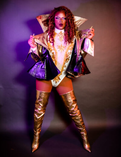 Aimee Yonce Shennel in a white and gold leotard and royal purple jacket, with gold thigh high boots.