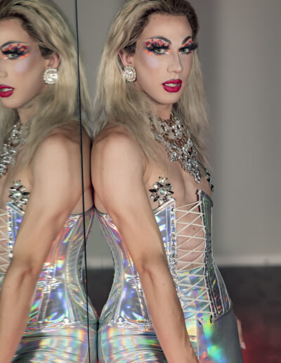Benz Menova wearing a silver holographic outfit, leaning against a mirror.