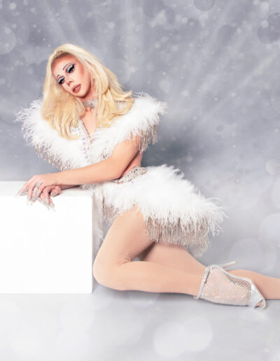 Benz Menova posing on a white block in a white feather outfit with silver fringe.