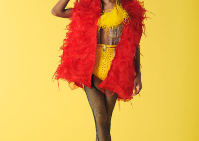 Karamilk wearing a yellow bikini with feathered top and red feather vest touching the crown on her head.