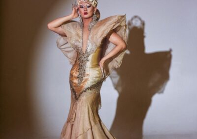 Lena Di posing in the spotlight wearing a champagne mermaid dress and champagne updo wig with tall silver and jeweled crown.