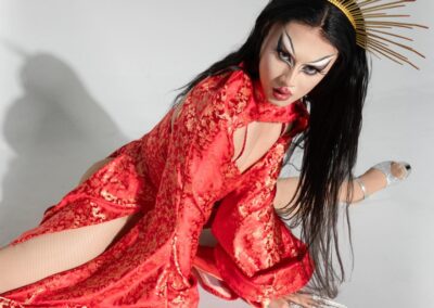 Lena Di in red silk dress and gold sun crown doing the splits.