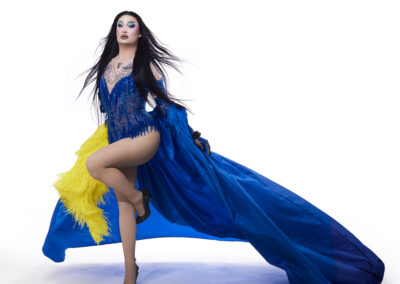 Van Goth posing dramatically wearing a blue rhinestone body suit, flourishing a shiny blue cape with a yellow feather accent.