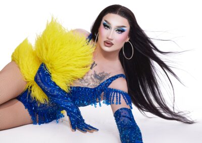 Van Goth posing on the floor, wearing a blue rhinestone corset with matching opera gloves, and a yellow feather accent on her right arm.