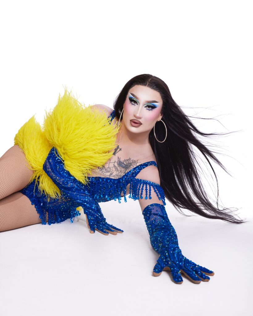 Van Goth posing on the floor, wearing a blue rhinestone corset with matching opera gloves, and a yellow feather accent on her right arm.