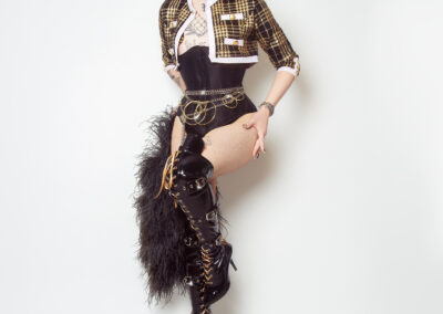 Van Goth in a black underbust corset, with black and gold plaid bolero jacket, gold accessories, thigh high boots, and feather boa.