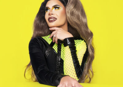 Vanity Affair posing at a yellow table wearing a black and yellow sleeved bodysuit.