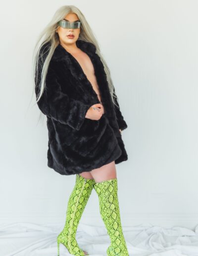 Chelazon Leroux posing to her left, wearing a black fur coat and green thigh high boots.