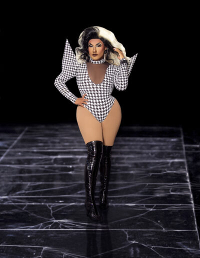 Dulce wearing a houndstooth bodysuit with dramatic triangular shoulders and black thigh high boots.
