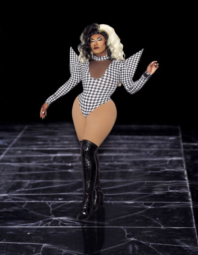 Dulce wearing a houndstooth bodysuit with dramatic triangular shoulders and black thigh high boots, posing like she's swaggering down a runway.