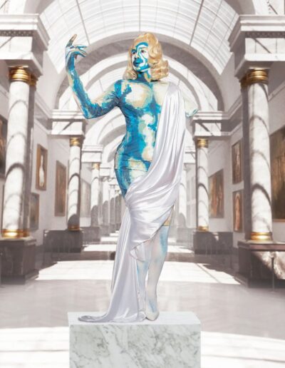 Halal Bae dressed and painted like a one-armed blue, white, and gold marble statue, posing with the remaining arm raised upwards.