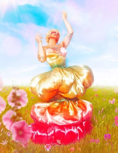 Halal Bae wearing a shiny, poofy, yellow and pink gown, posing dramatically with her arms raised toward the sky.