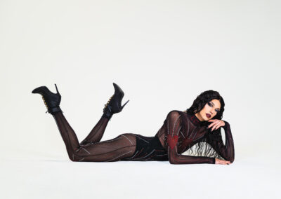Liquorice wearing a sheer rhinestoned bodysuit over a black underbust corset, lying on the ground and propping herself up with her left arm.