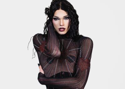 Hips up view of Liquorice wearing a sheer rhinestoned bodysuit over a black underbust corset, holding her waist with her left arm and her neck with her right arm.