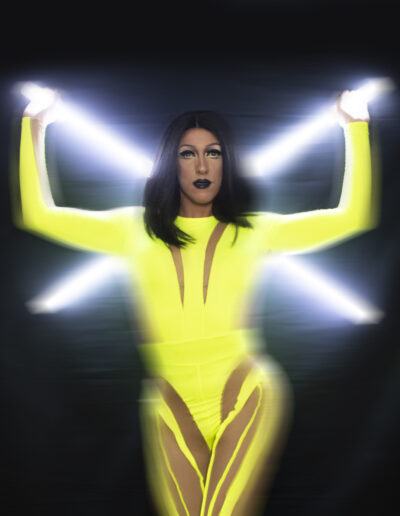 Mya Foxx in a bright yellow bodysuit, holding two white light sticks in an X behind her back. The image is blurred around her body.