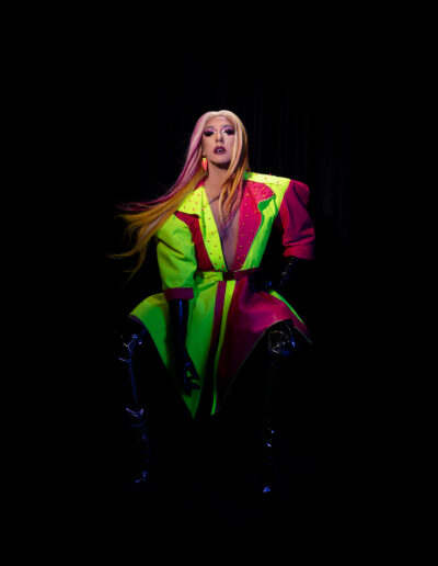 Mya Foxx in a neon pink and green jacket with black thigh high boots, posing in a chair against a black background.