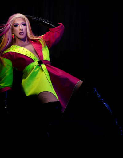 Mya Foxx in a neon pink and green jacket with black thigh high boots, leaning back in a chair against a black background.