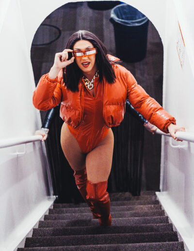 Mya Foxx in an orange leotard and matching orange puffer jacket and thigh high boots, walking up a flight of stairs while touching her shades.