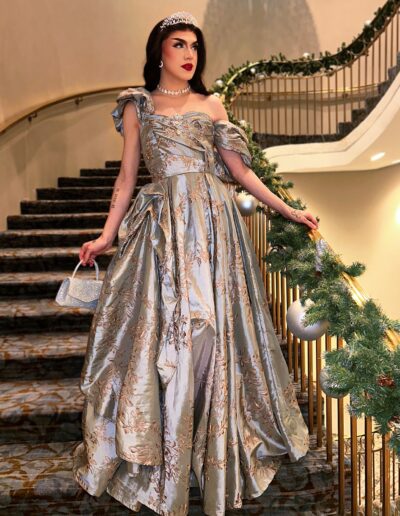 Nicki Nastasia wearing a silver and gold-embroidered evening gown, walking down a curved staircase.