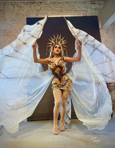 Nicki Nastasia wearing a gold corset, sun headdress, and white sheer flowing cape. She's holding the cape with her arms to look like wings.