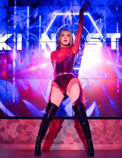 Nicki Nastasia performing on stage in a red leotard with fringe accents, posing with her left arm in the air.