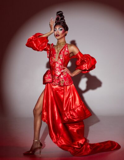 Phoenix Black wearing a red silk gown with puffy detached sleeves, posing with her left hand on her hip and right hand against her temple.