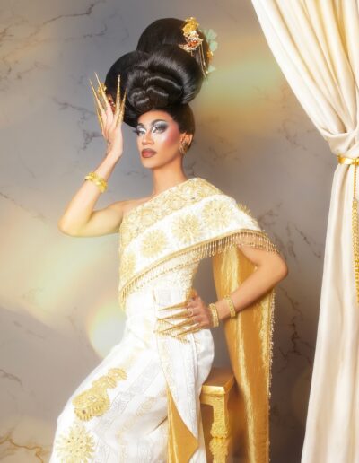 Phoenix Black wearing a white and gold silk gown, seated on a stool with her left hand on her hip and right hand next to her face.