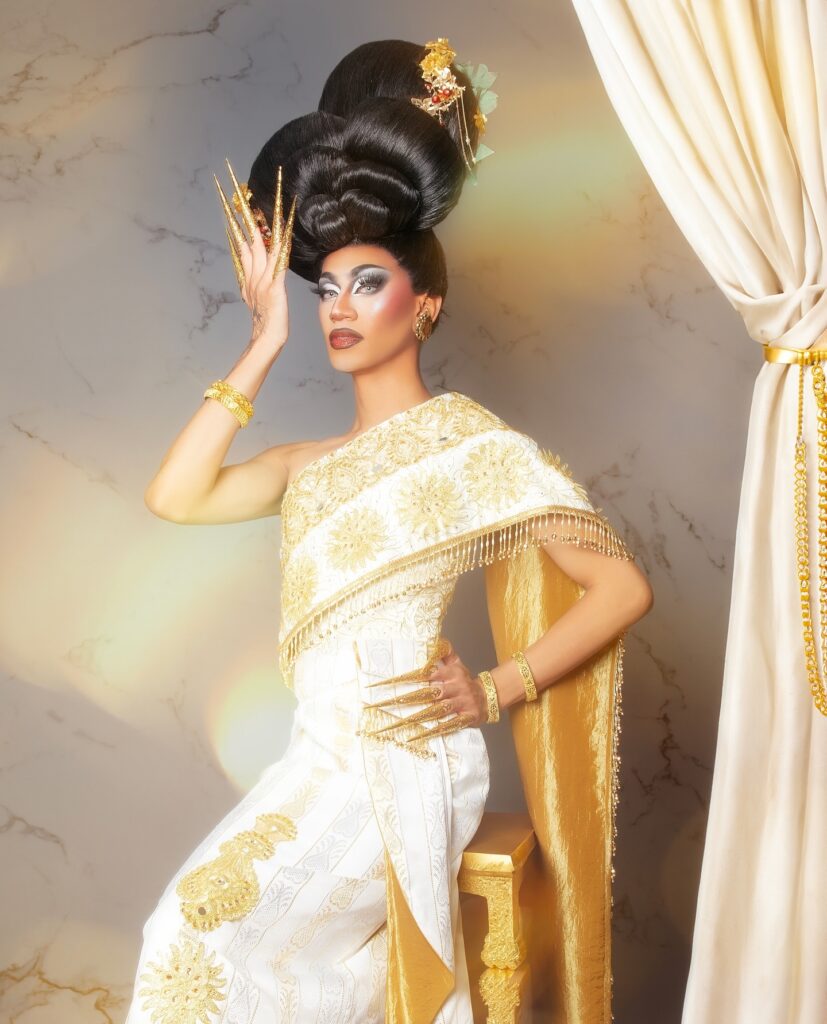 Phoenix Black wearing a white and gold silk gown, seated on a stool with her left hand on her hip and right hand next to her face.