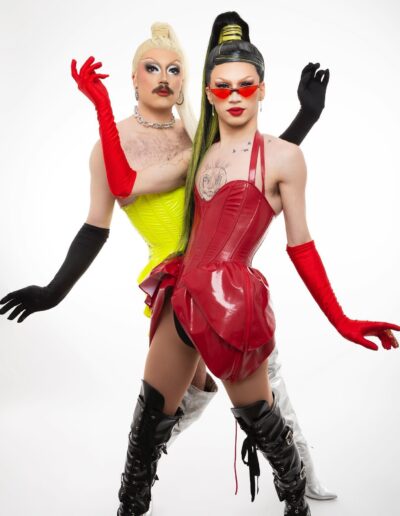 Rosie in a Red Leather corset with black thigh high boots and red opera gloves, posing symmetrically with Logan the Queen wearing a yellow corset and black opera gloves.