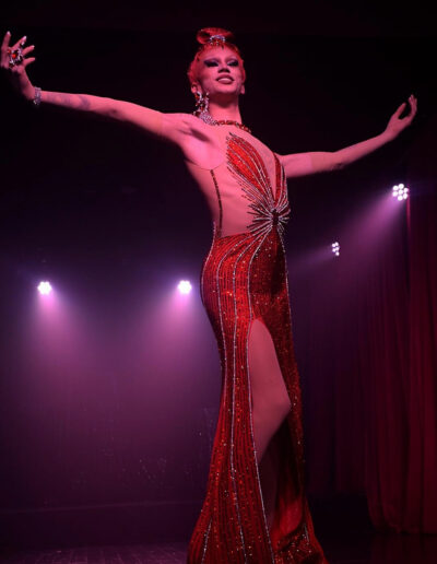 Low view of Rosie in a red rhinestone gown, arms out and posing on a stage.