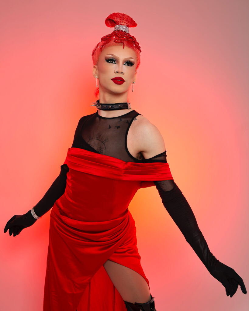 Rosie in a red silk dress over a black sheer tank top with matching black opera gloves, posing dramatically.