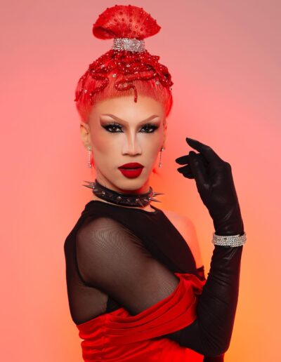 Rosie in a red silk dress over a black sheer tank top with matching black opera gloves, gazing intently to the camera with her right arm lifted by her face.