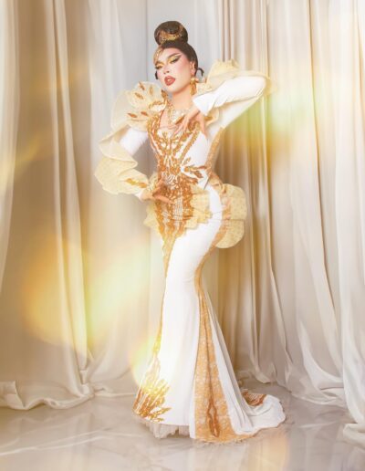 Star posing with her left arm raised and right arm lifted slightly, wearing a white and gold embellished gown.