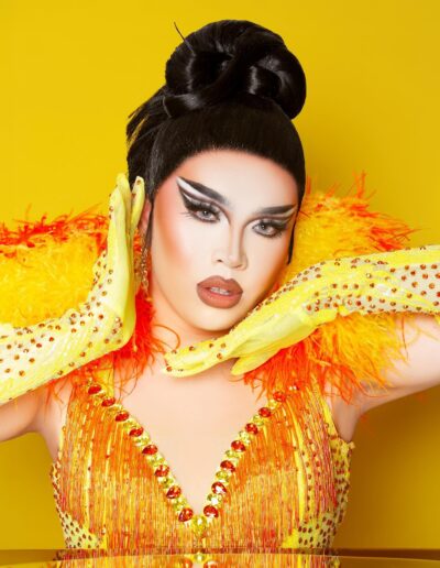 Star wearing a yellow corset with red accents and feathered collar with matching yellow rhinestone gloves. She's framing her face with her hands.