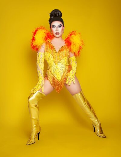 Star wearing a yellow corset with red accents and feathered collar with matching yellow rhinestone gloves and gold thigh high boots. She's leaning forward on her right leg, with her left hand on her hip and left leg extended back slightly.