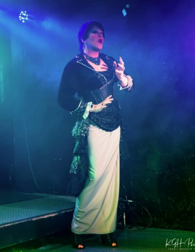 Geena Hayze performing in a white and black floor length dress with black blazer, hip shawl, and black corset belt.