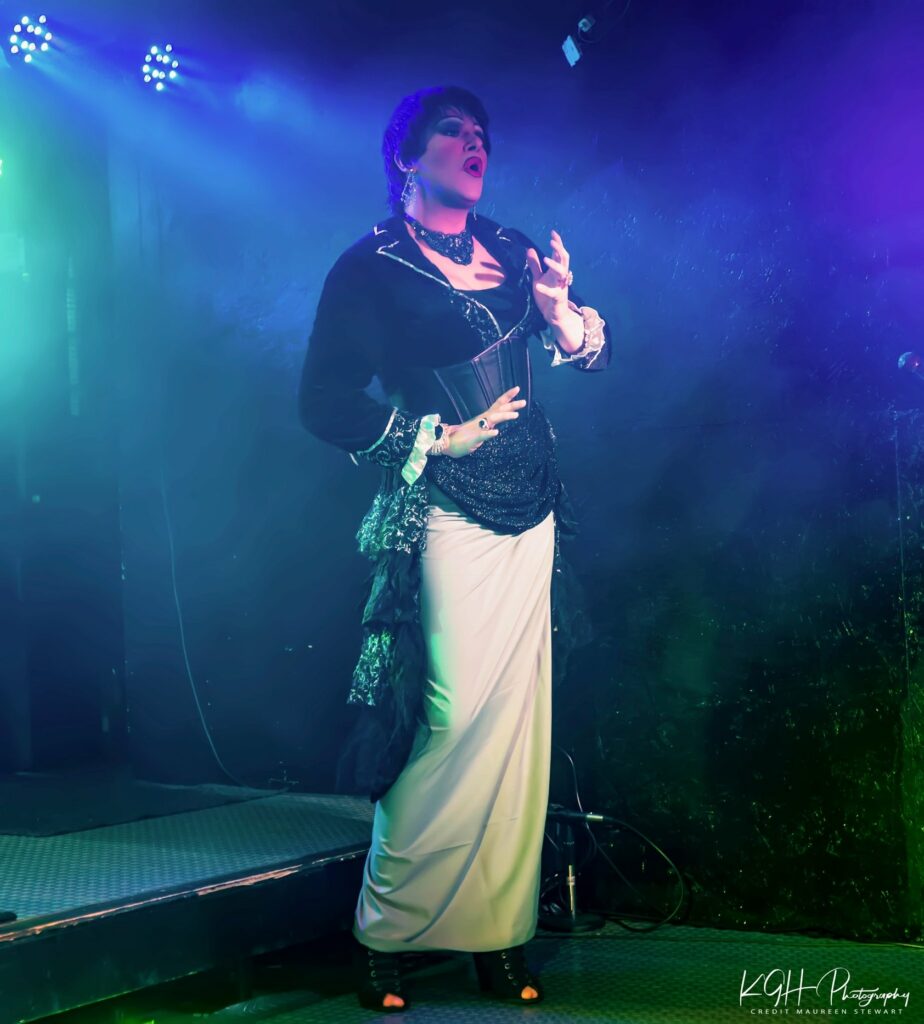 Geena Hayze performing in a white and black floor length dress with black blazer, hip shawl, and black corset belt.