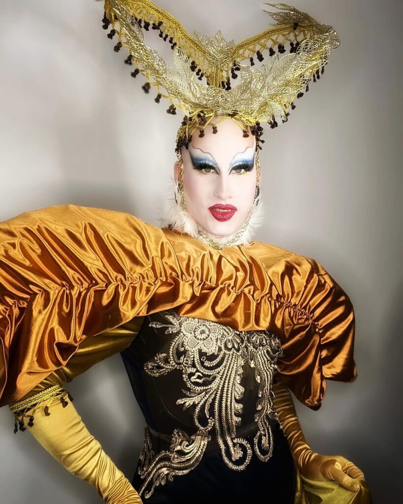 Lua wearing a dark corset with gold embroidery and shiny bronze puffy sleeves, topped with an elaborate gold headdress with black bead accents.