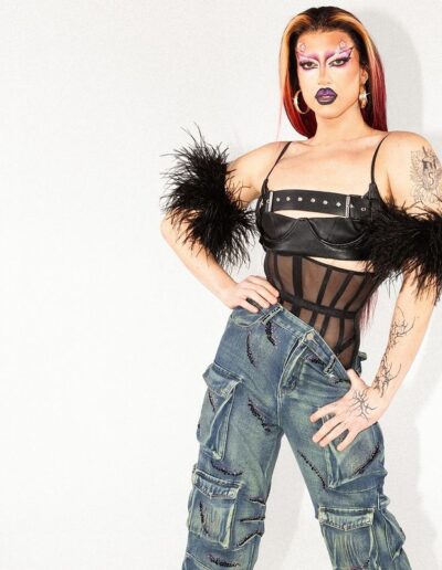 Saltina Shaker wearing a black sheer underbust corset, black strappy crop top with feather armbands, and grey denim jeans. She's posing with her right arm on her hip and her left arm tugging the waistband of the jeans down with her thumb.